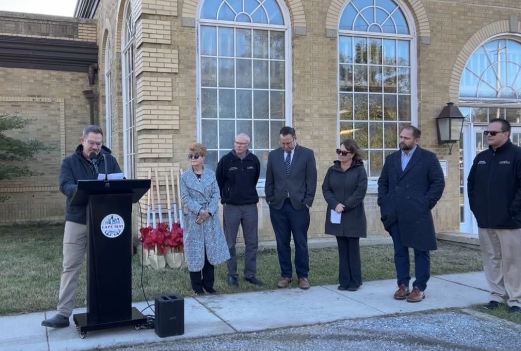 A speaker at a City of Cape May podium addresses the groundbreaking as six officials stand in front of the 1926 Cape May Water Works building, beside a row of ceremonial gold shovels with red bows