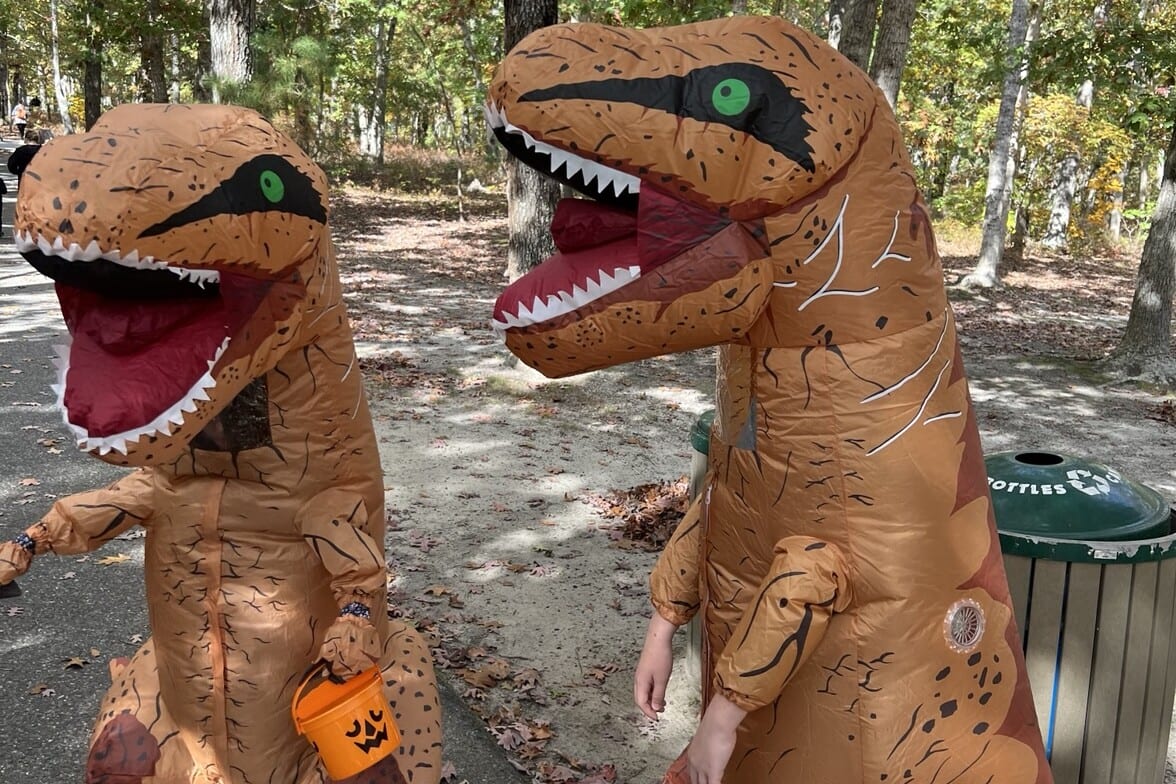 Two children in inflatable T. rex costumes, one carrying a jack-o'-lantern bucket, on a wooded path at the Cape May County Park and Zoo